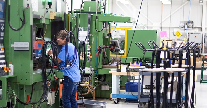 A Interpower worker in a blue shirt and safety glasses operates a large green industrial power cord molding machine in a clean, well-lit manufacturing facility. The machine has various cables and control panels, and the worker is focused on the task inside the machine's open panel. Nearby, coiled black cables hang from racks, and other equipment and workstations are visible in the background. The environment suggests a high-tech or precision manufacturing setting.