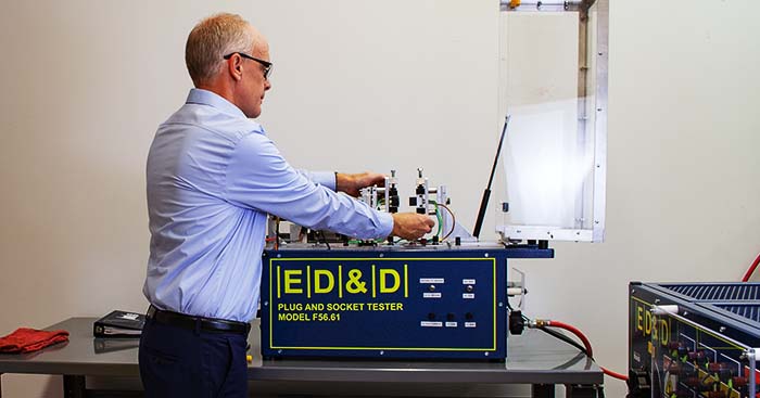 This image shows Ron Barnet, Product Development Specialist, operating an ED&D Plug and Socket Tester Model F56.61, a specialized piece of testing equipment. Ron is wearing a formal light-blue shirt and safety glasses, focused on the task. The machine, designed for plug and socket testing, has several wires, switches, and connectors visible, showing it's being used for electrical testing and quality assurance purposes.  The environment is Interpower's testing or lab, and there is another device of similar branding visible on the right. The workspace is organized, with a clean bench and a book or manual nearby. The presence of a safety screen (transparent cover) on the equipment highlights a focus on safety while testing electrical components.